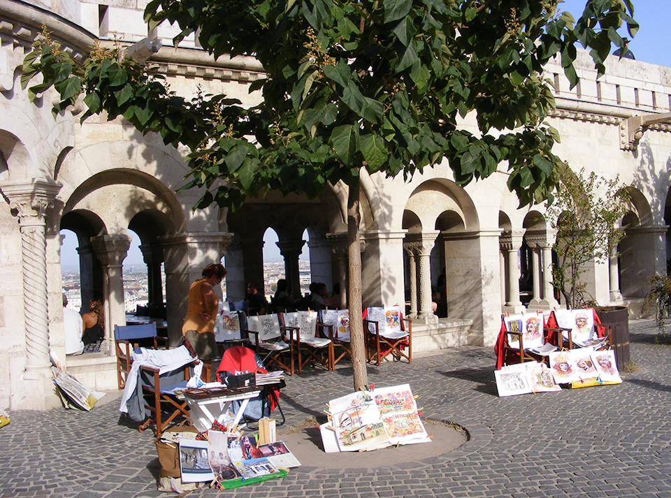 Outdoor art market with paintings displayed under a tree. Arched building in the background with people sitting inside.