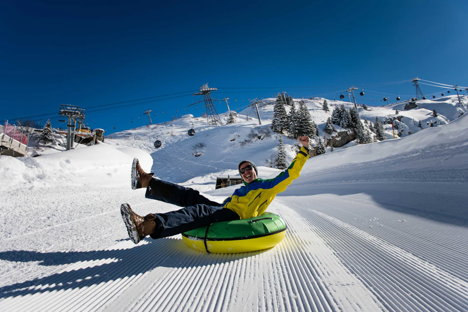 A person in winter gear rides a yellow and green snow tube down a groomed snow slope under a clear blue sky.