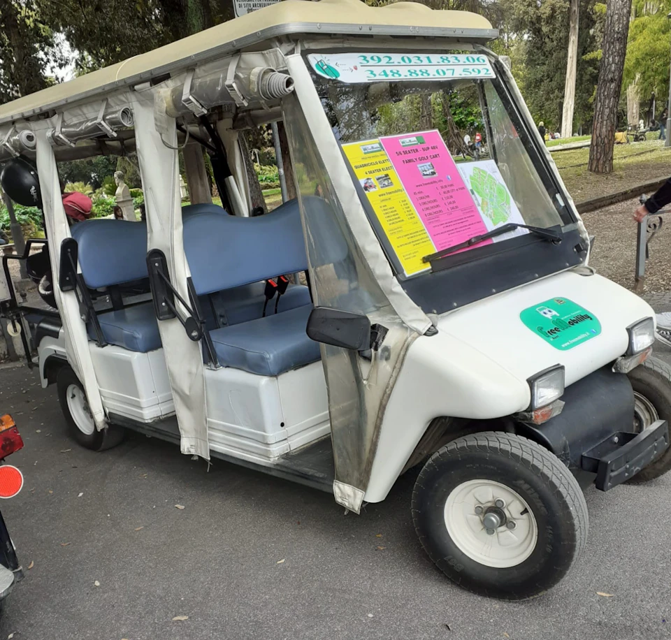 White electric cart with blue seats and advertisements on the windshield, parked on a paved path in a park setting.