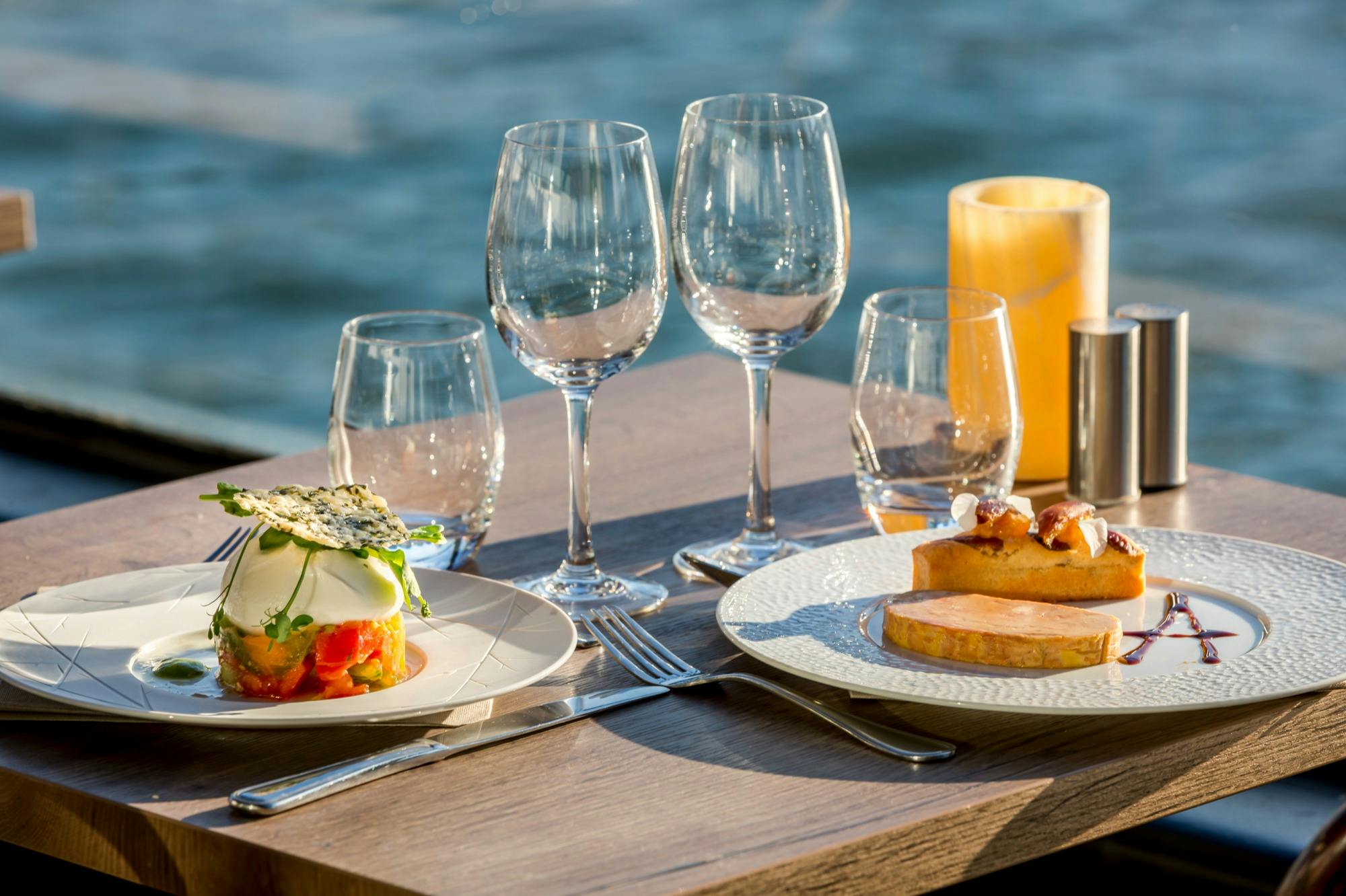 Zwei Teller und vier Weingläser auf einem Holztisch im Freien mit einer Kerze, vor einem Hintergrund am Wasser.