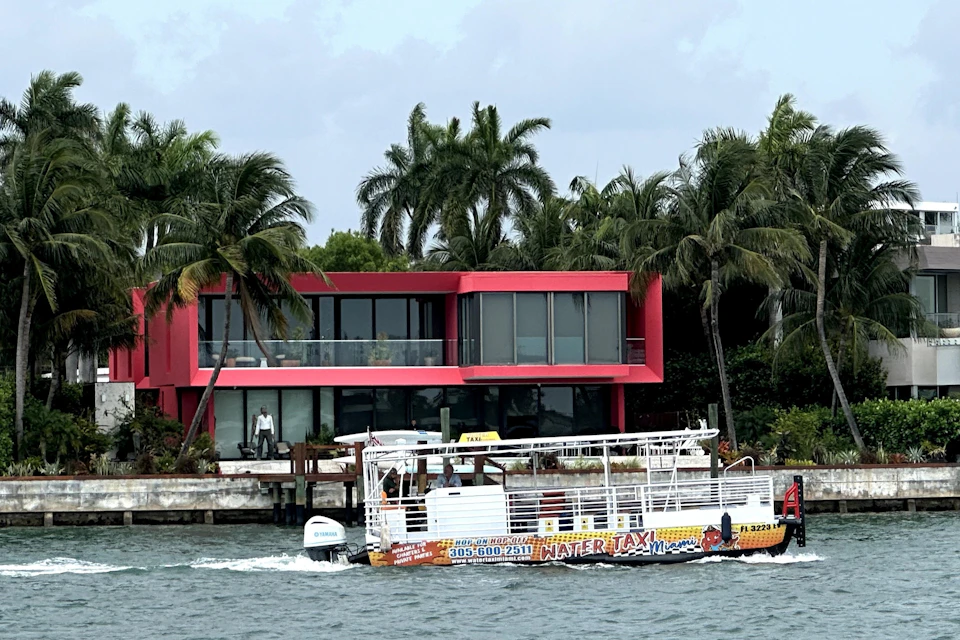 A pink modern house with large glass windows by the water, surrounded by palm trees, with a water taxi passing in front.