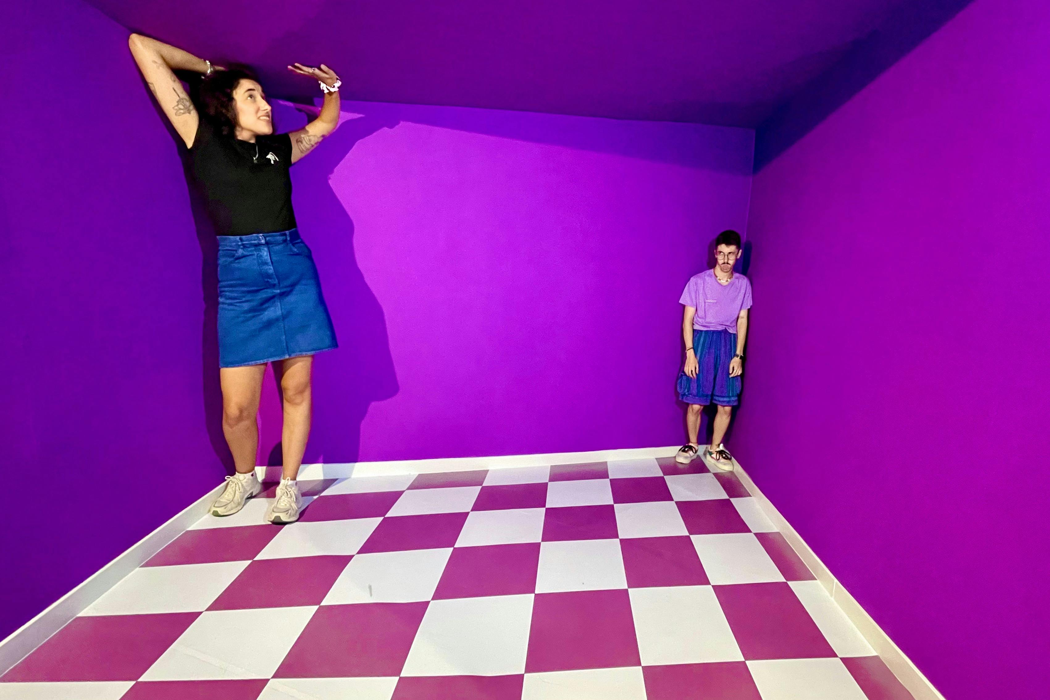 Una persona està parada en un racó d'una vibrant habitació de color porpra amb terra a quadres de color rosa i blanc, vestida amb roba informal.