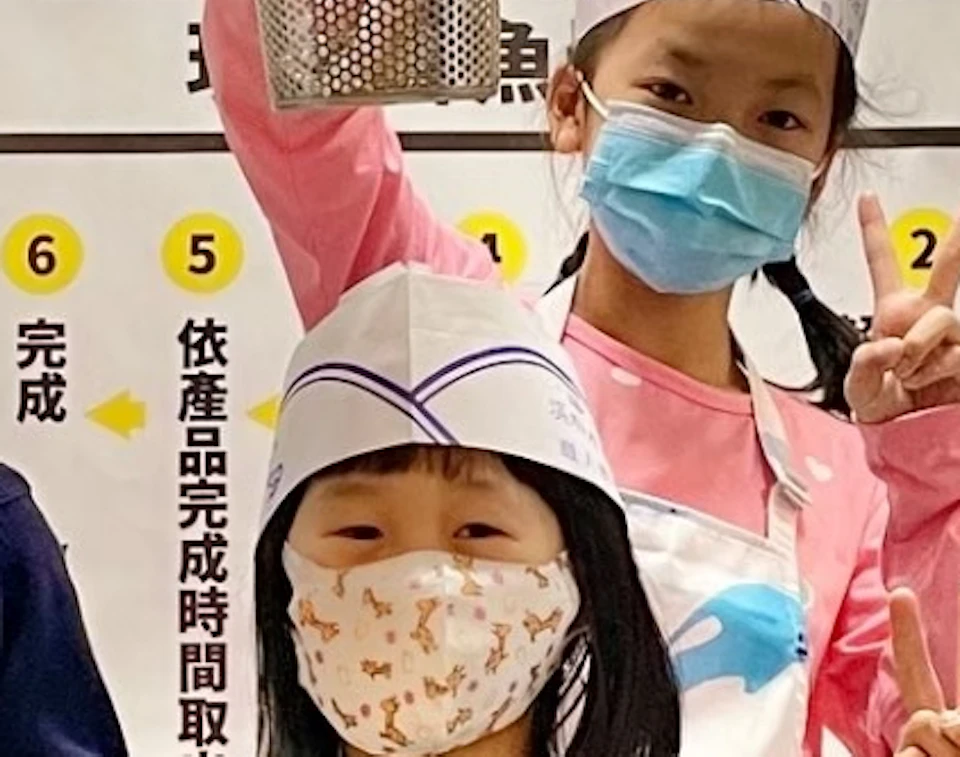 Five children wearing aprons and hats stand in front of a safety instructions board, holding metal baskets and smiling.