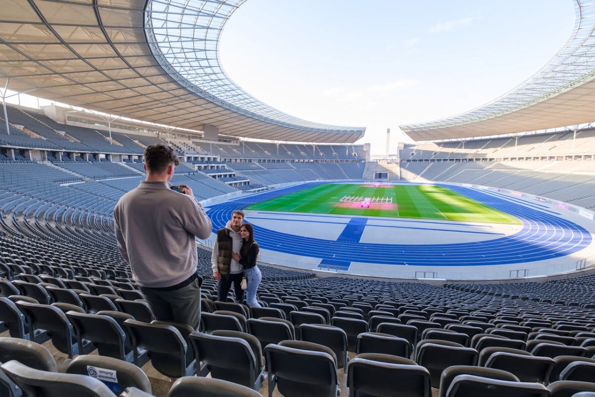 Eine Person macht ein Foto von einem Paar in einem großen, leeren Stadion mit blauen Schienen und einem grünen Feld im Hintergrund.