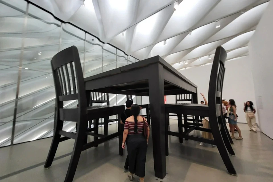 People walking under an oversized table and chairs in a modern exhibition space with a high, white ceiling.