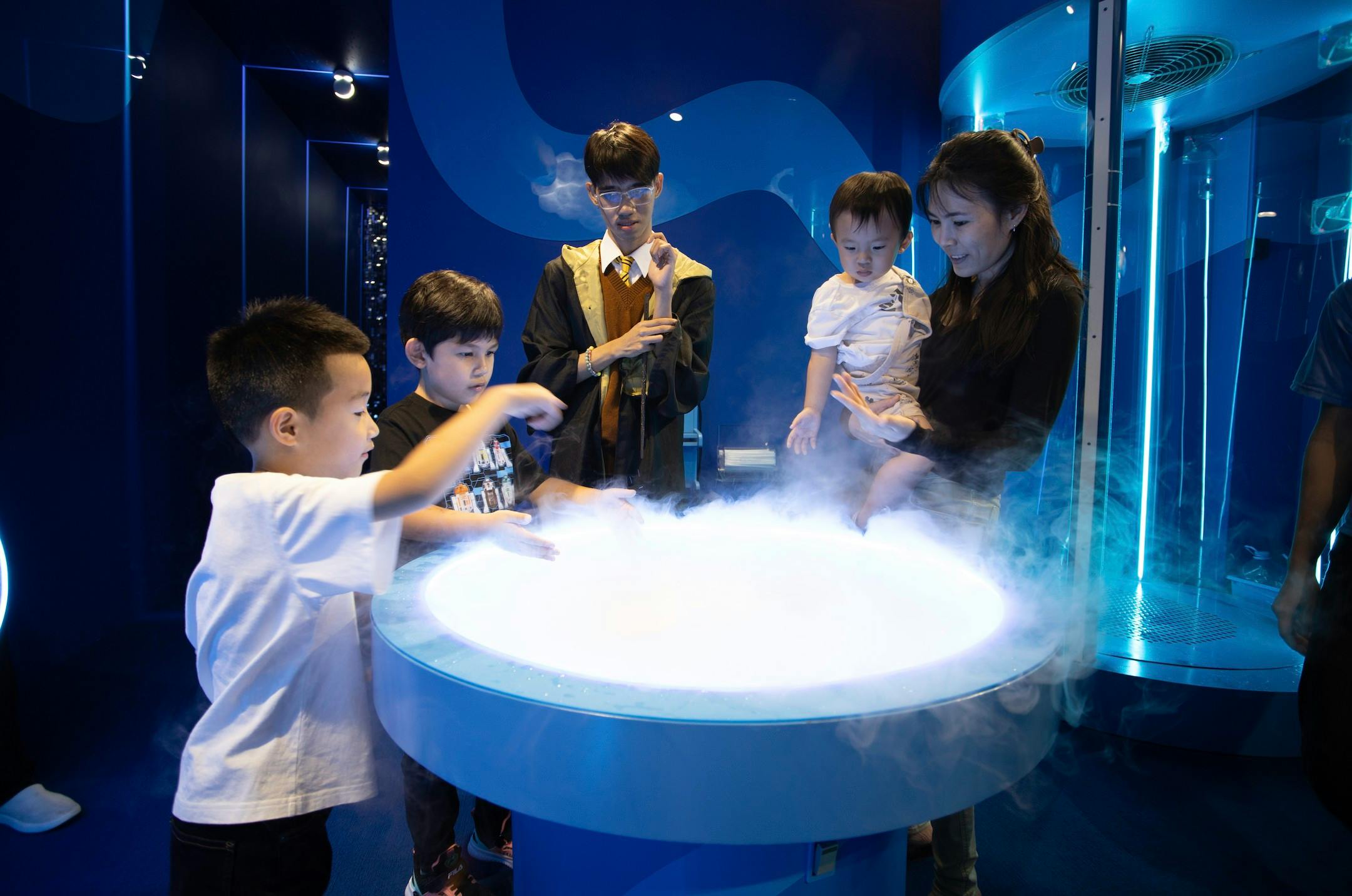 A group of people, including children and an adult, interact with a misty display table emitting fog in a dimly lit room.