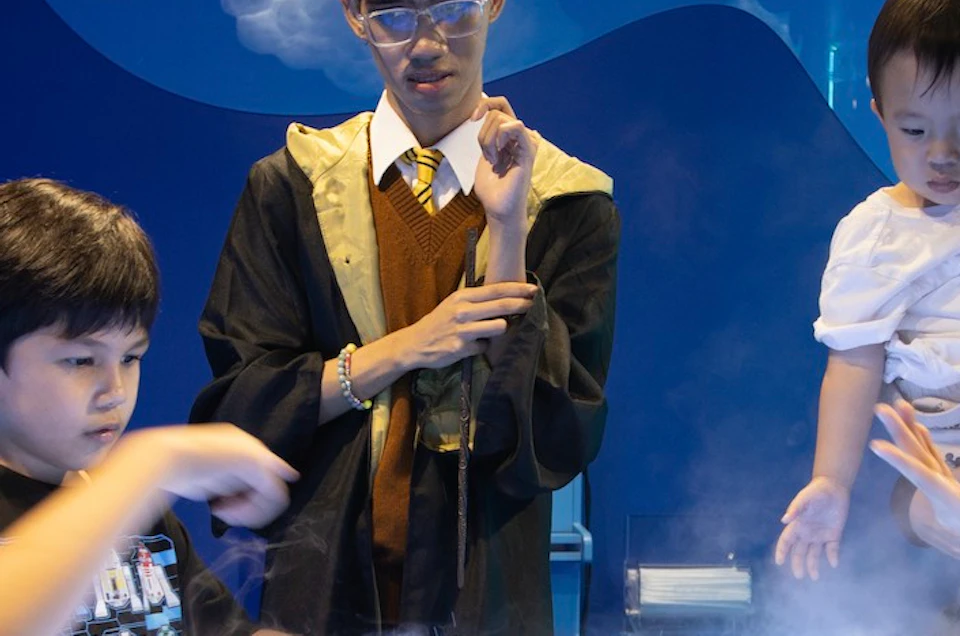 A group of children and adults interact with a mist-covered circular display in a blue-lit room.