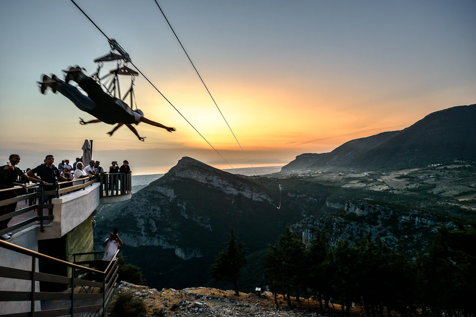 Person zip-lining over a mountainous landscape at sunset with observers on a platform.
