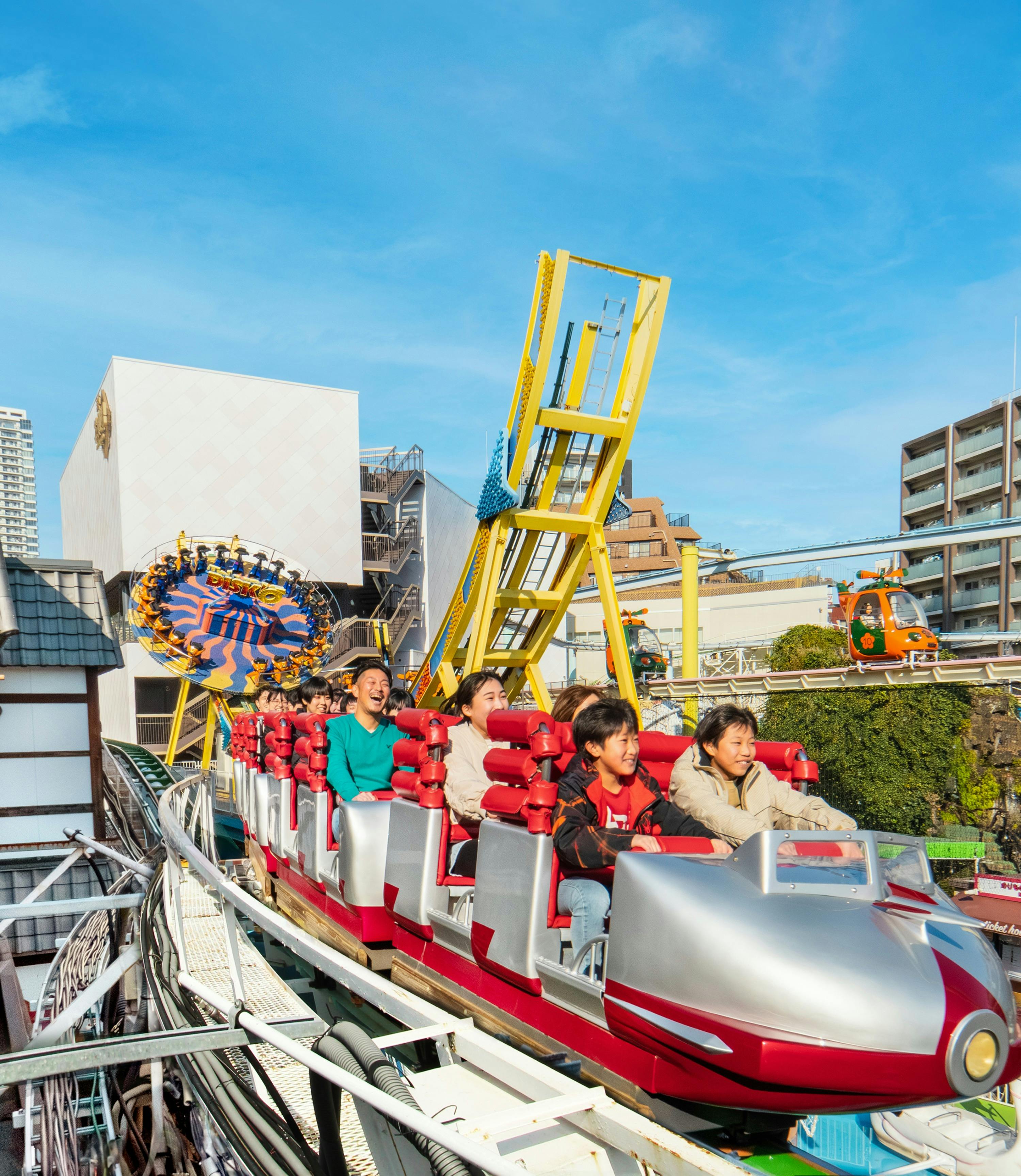 People riding a roller coaster with red and silver cars in an amusement park, with other attractions visible in the background.