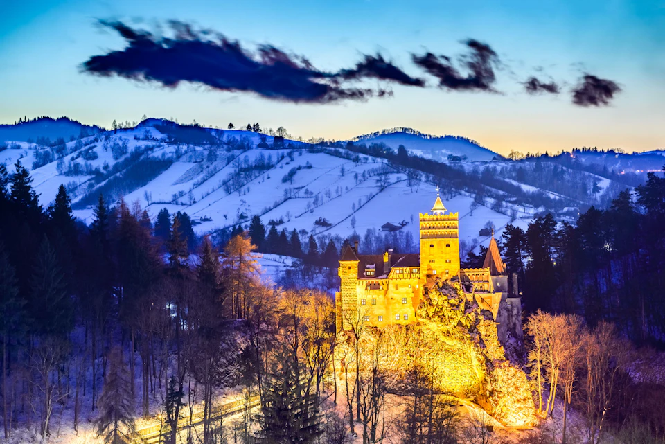 A brightly lit castle at dusk, surrounded by snow-covered mountains and trees under a twilight sky with dark clouds.