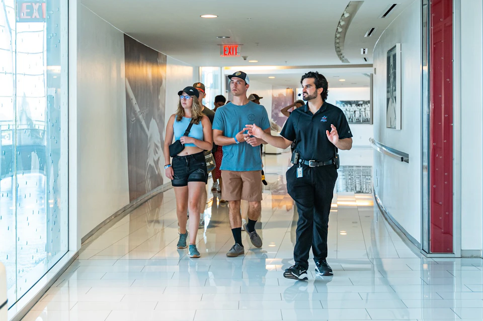 A guide gestures while leading a group of visitors through a bright hallway with glossy flooring and wall decor.
