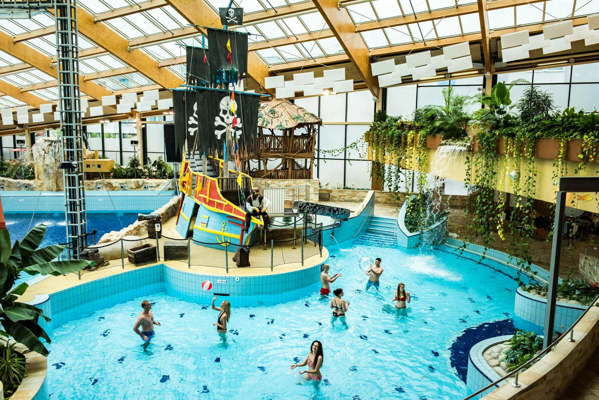 Indoor water park with pirate ship, waterfall, and people swimming and playing in the pool beneath a wooden roof structure.