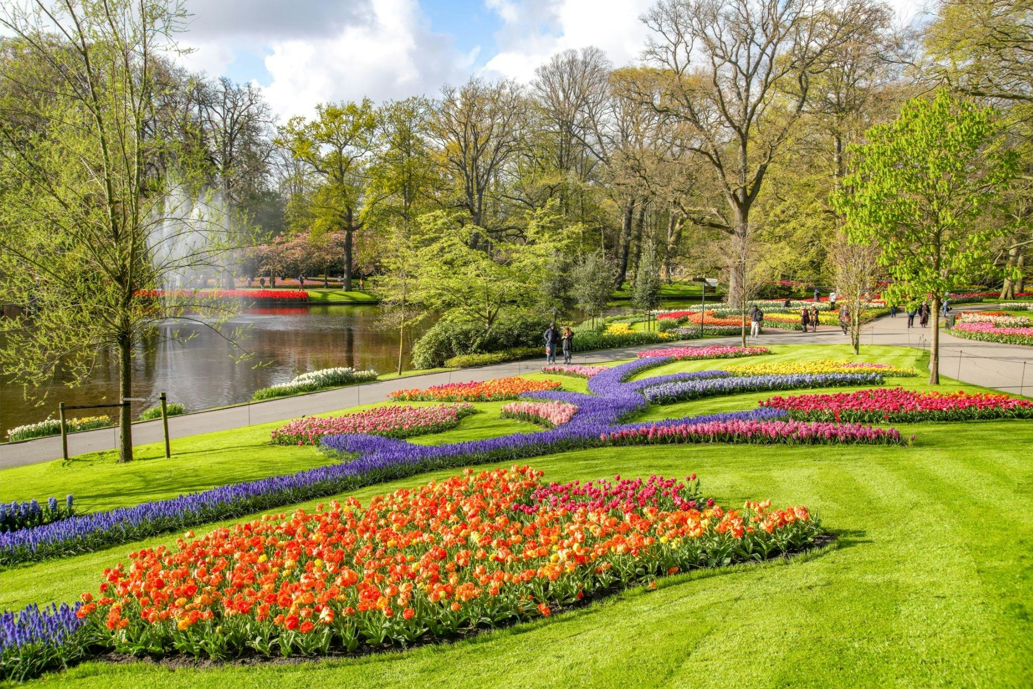 A vibrant garden with colorful flower beds, green lawns, trees, a pond, and people walking along a path under a cloudy sky.