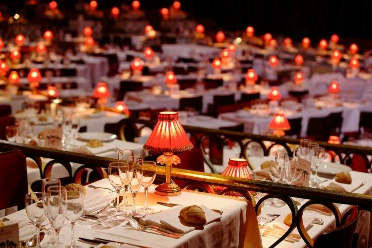 A restaurant with tables set with white cloths, red lamps, glassware, and bread, creating a warm and elegant atmosphere.
