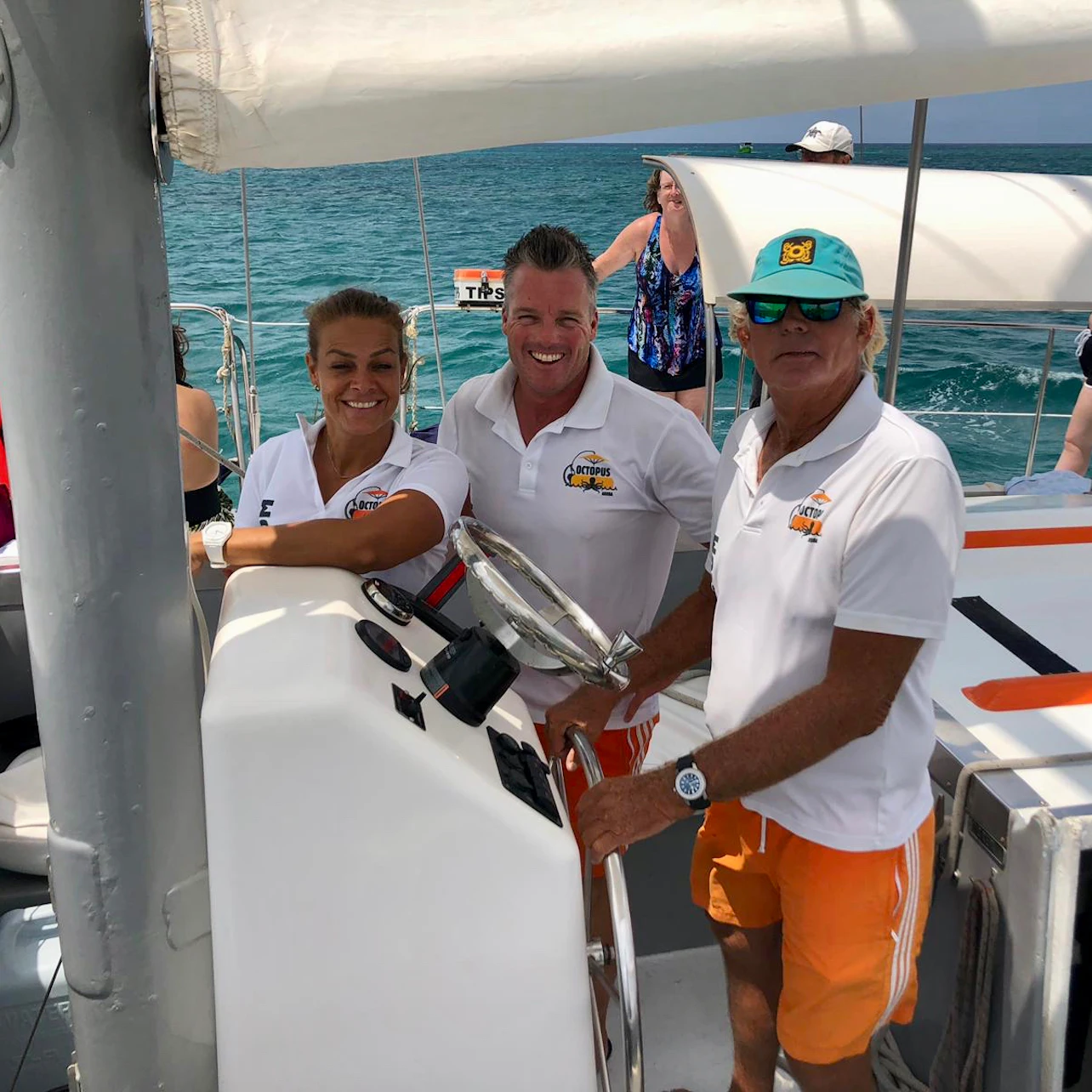 Three people in matching attire stand aboard a boat, smiling at the camera with the ocean in the background.