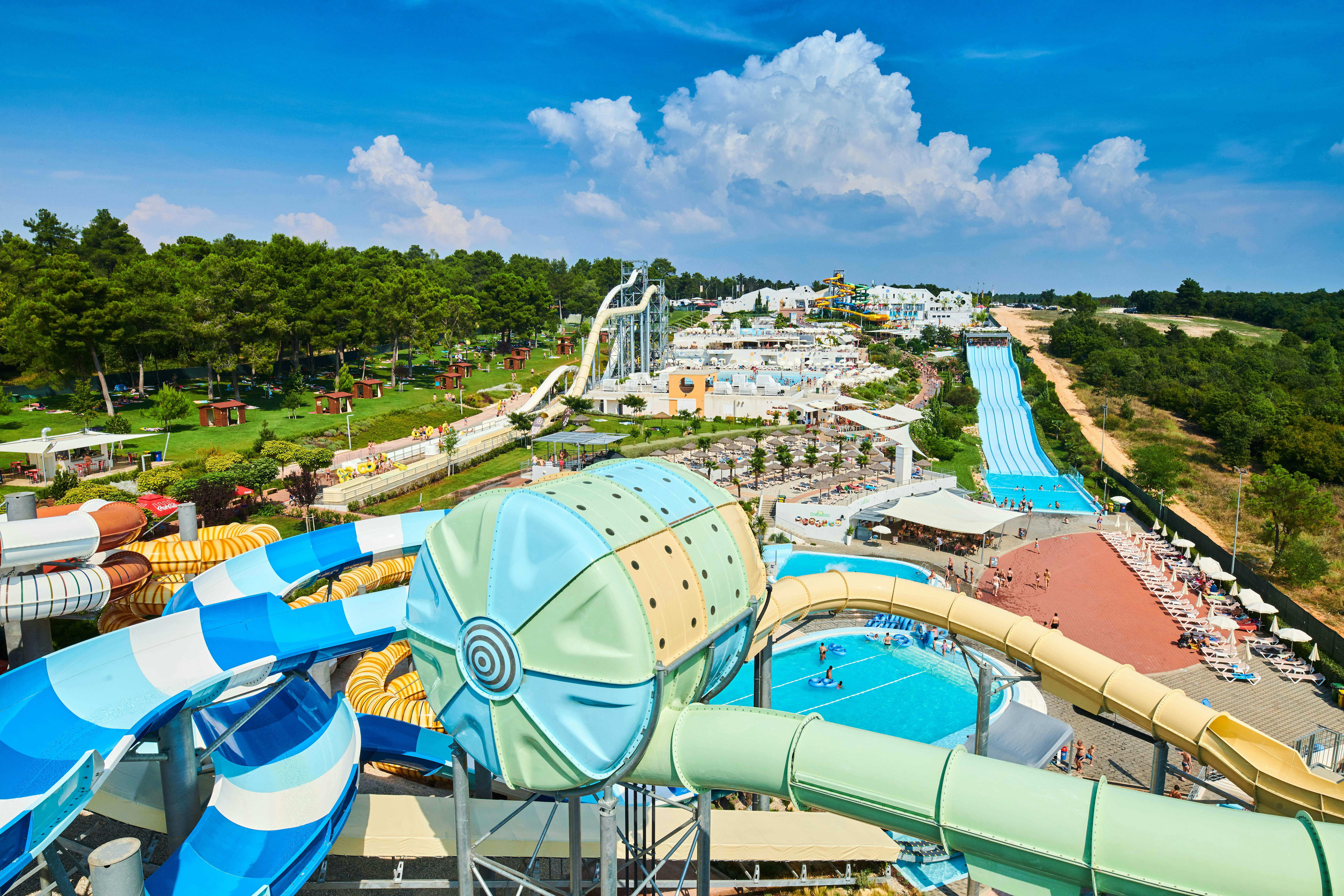 Luftaufnahme eines Wasserparks mit verschiedenen Rutschen, Becken, grünen Bäumen und klarem blauen Himmel.