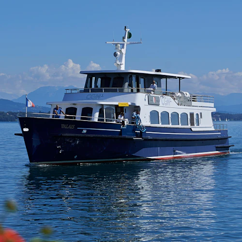 En blåvit passagerarbåt som heter "Valais" navigerar en lugn sjö under en klar himmel, med berg i bakgrunden.