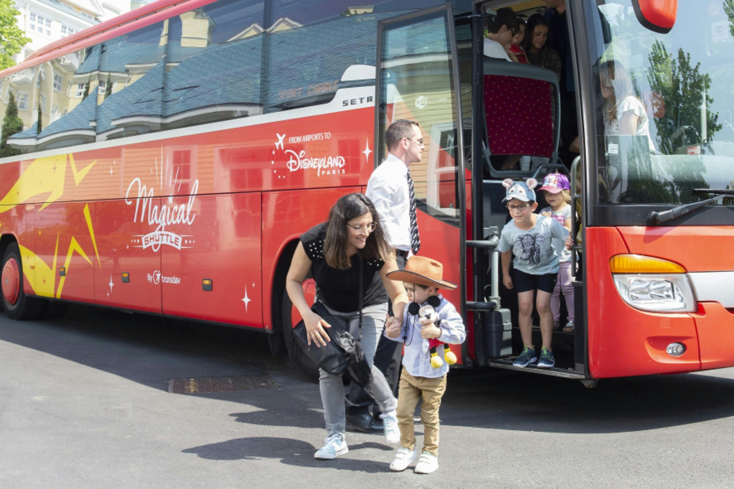 Children and a woman exit a red bus with a Disneyland Paris logo. A boy in a cowboy hat holds a Mickey Mouse plush toy.