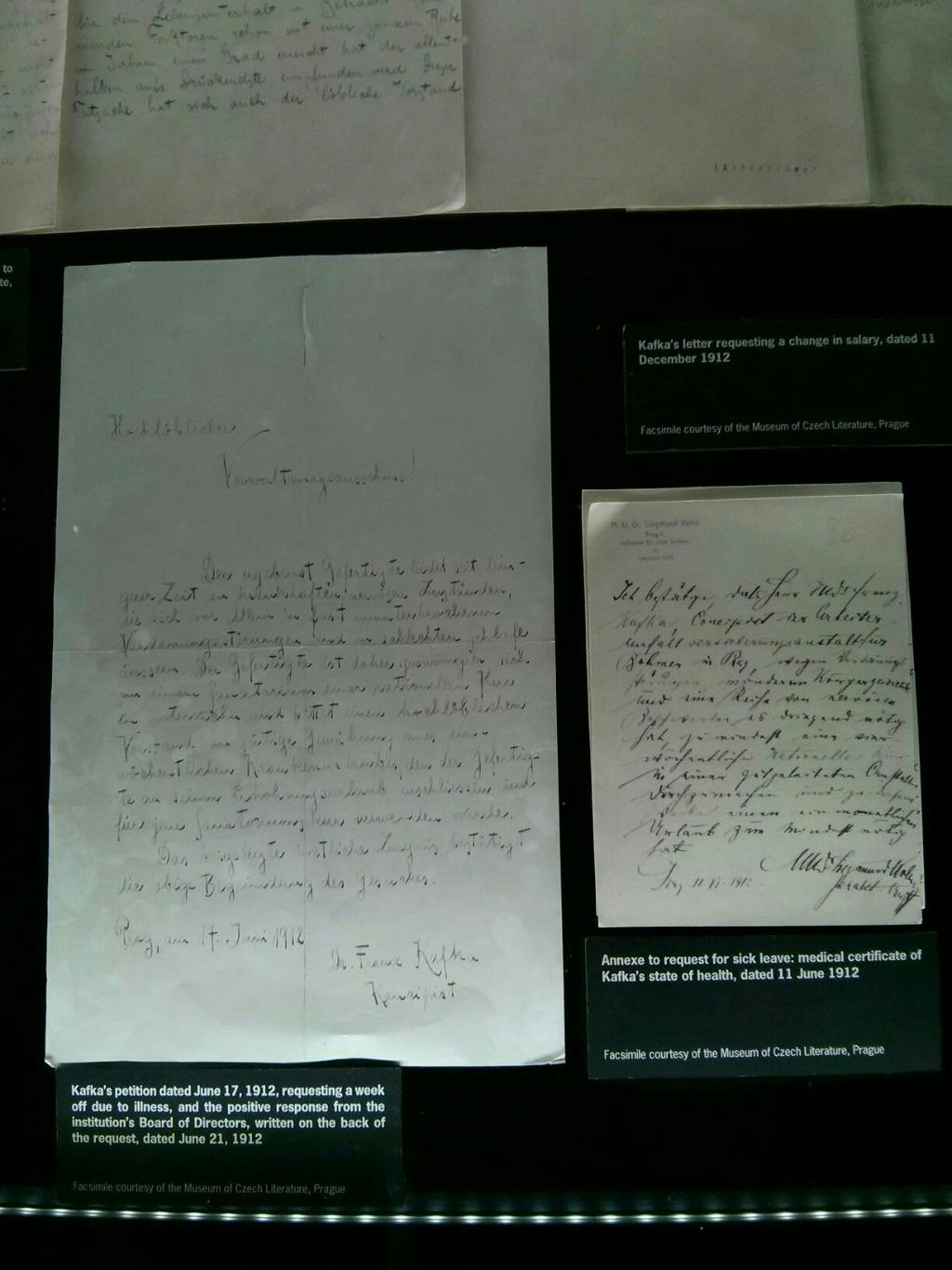 Documents manuscrits affichés sur un fond noir avec des étiquettes explicatives indiquant le contexte de Franz Kafka.