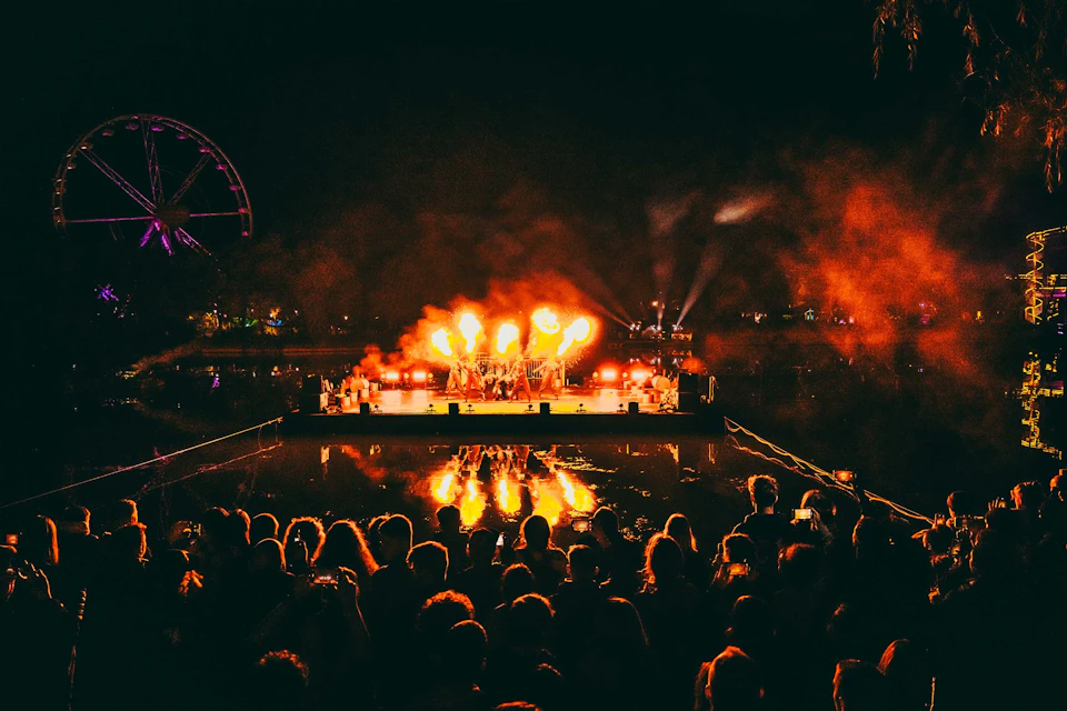 A nighttime outdoor concert with fire effects on stage, viewed by a crowd with a ferris wheel in the background.