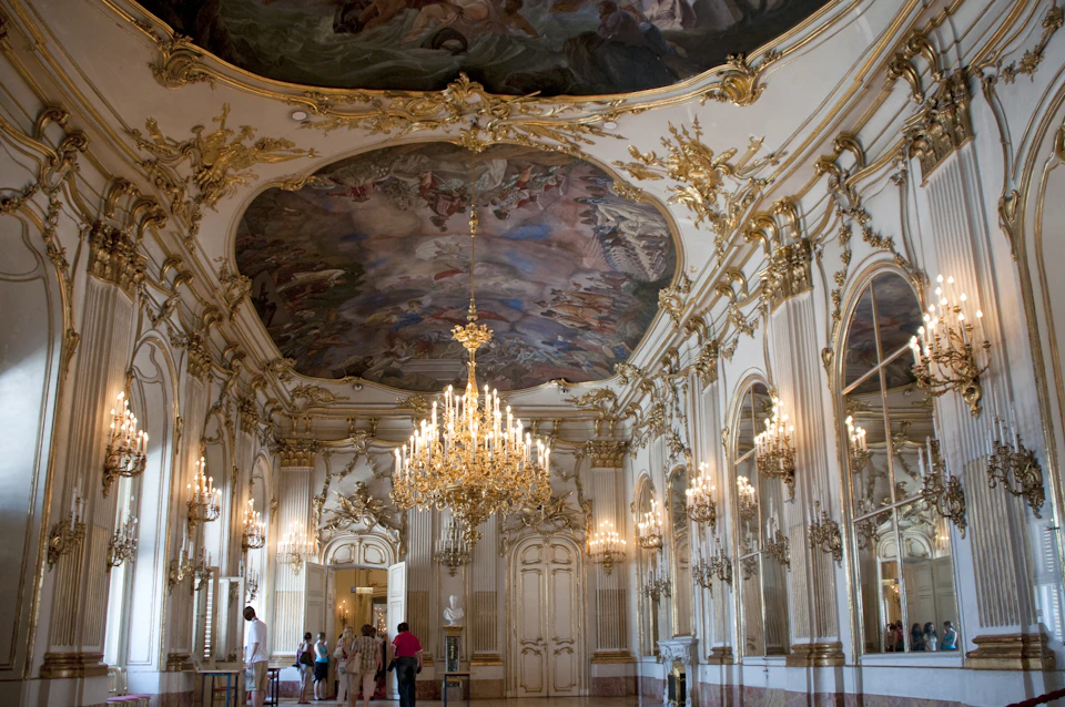 Ornate room with a large chandelier, gilded decorations, and a painted ceiling. Visitors in casual clothing admire the interior.