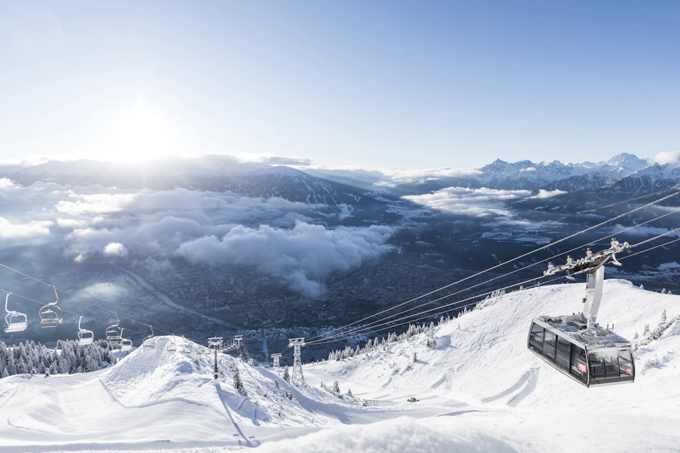 Snow-covered mountain landscape with a cable car, a sunlit sky, and clouds hovering over a valley below.