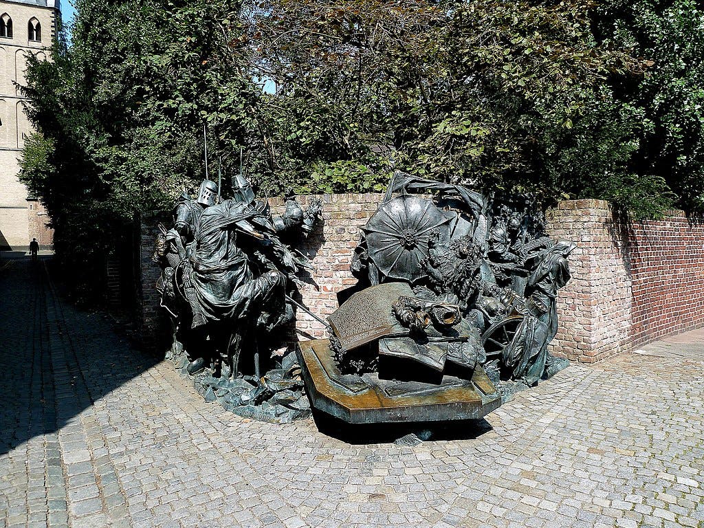 Bronze statue group depicting medieval knights and figures, set against a brick wall and surrounded by trees. Cobblestone path leads to it.