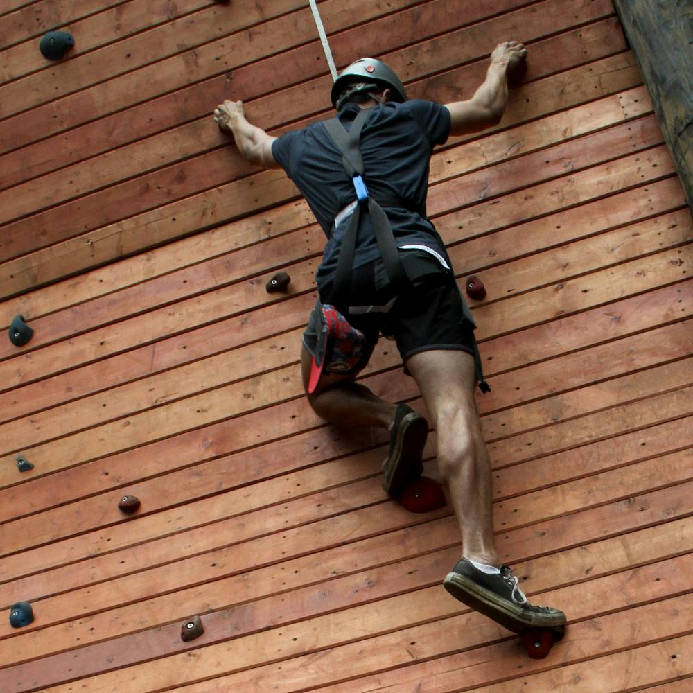 하네스, 헬멧, 검은색 반바지를 착용한 채 나무 벽을 등반하는 사람. 왼쪽 다리가 구부러져 다음 홀드를 향해 뻗습니다.