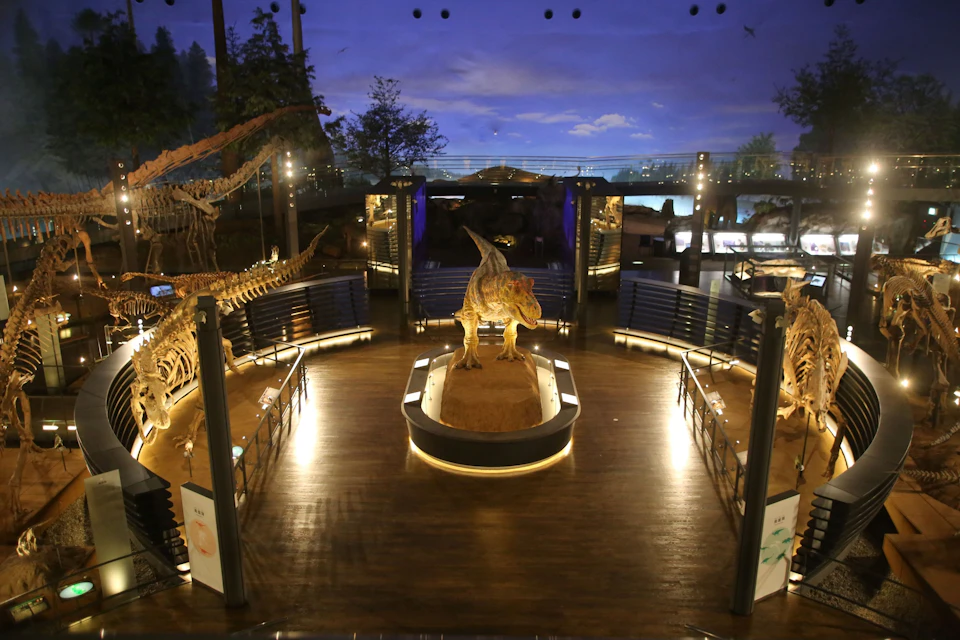A museum display featuring dinosaur skeletons and models, with a Tyrannosaurus rex centerpiece under dramatic lighting.