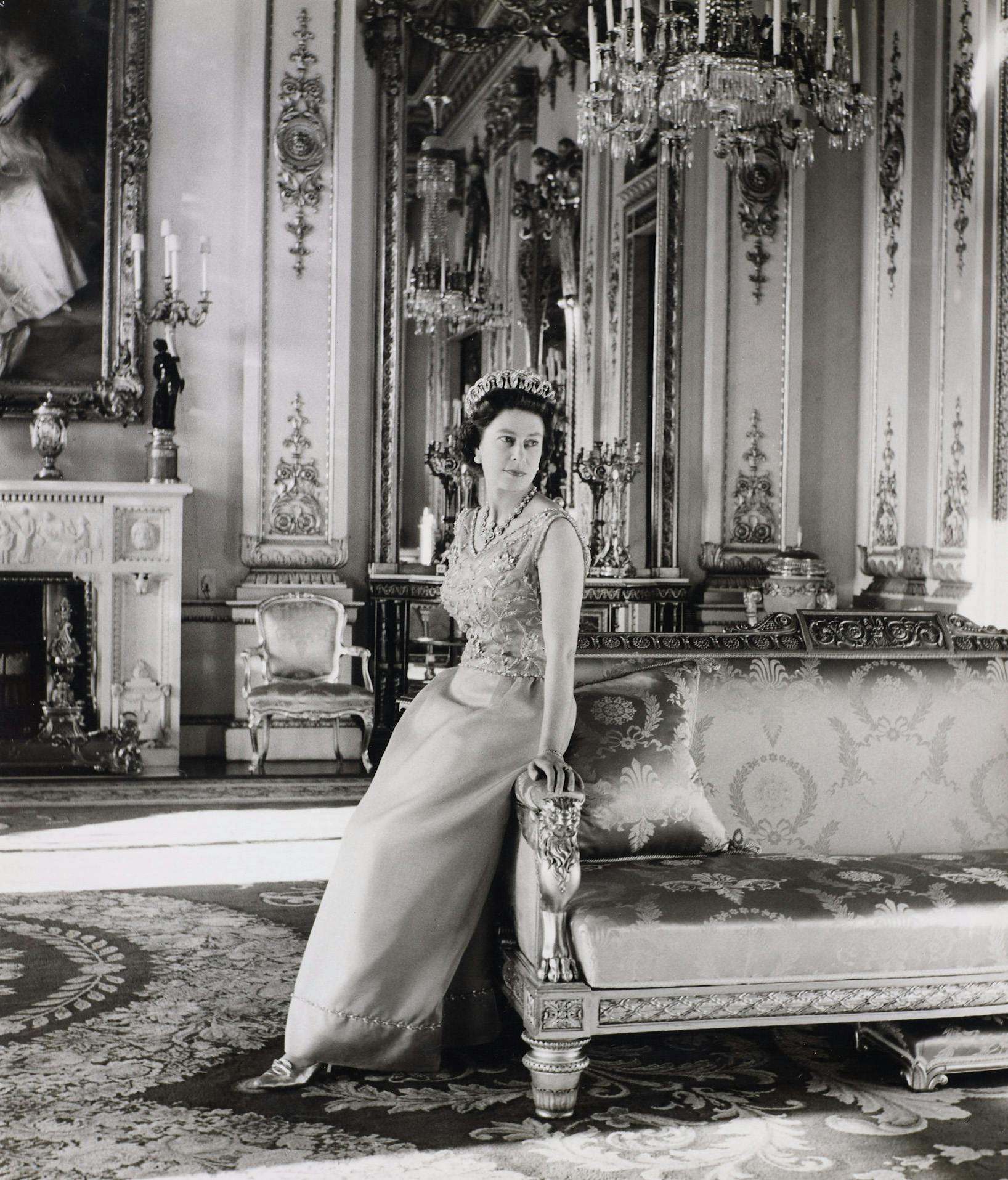 Una donna con diadema e abito da sera si appoggia su un divano decorato in una stanza elaborata con lampadari, opere d'arte e un camino.