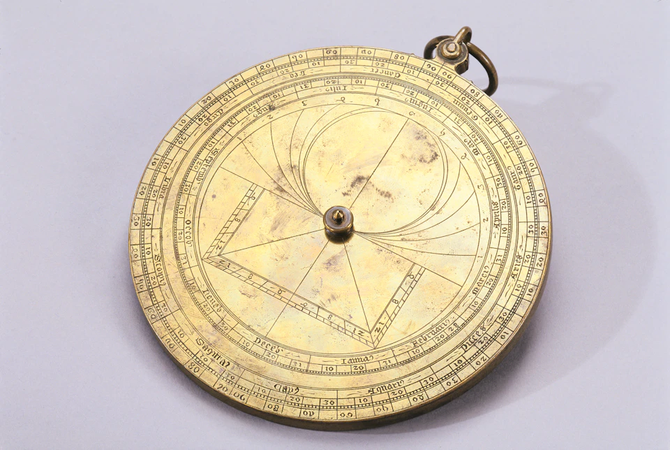 A brass astrolabe with intricate circular markings and geometric engravings; a small ring is attached to the top.