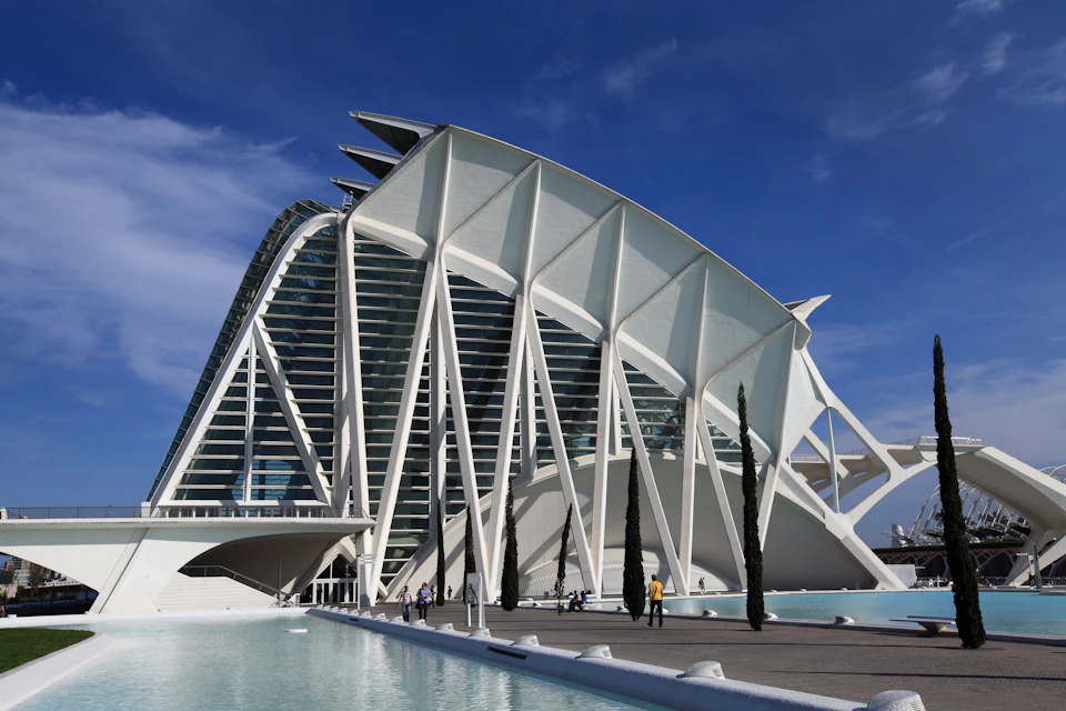 White, futuristic building with tall, angular structures and arched supports, surrounded by water and a few trees.