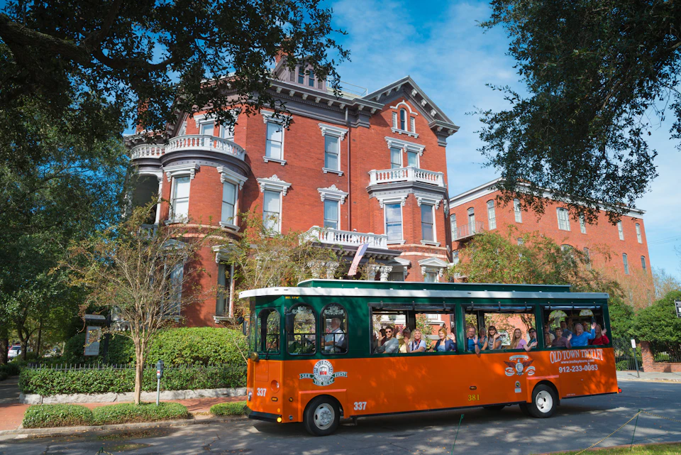 Group of tourists riding an orange and green trolley in front of a large, historic red-brick building with white trim.