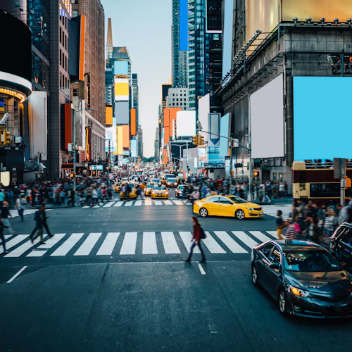 Belebte Stadtkreuzung mit starkem Fußgänger- und Fahrzeugverkehr, umgeben von hohen Gebäuden und Leuchtreklamen.