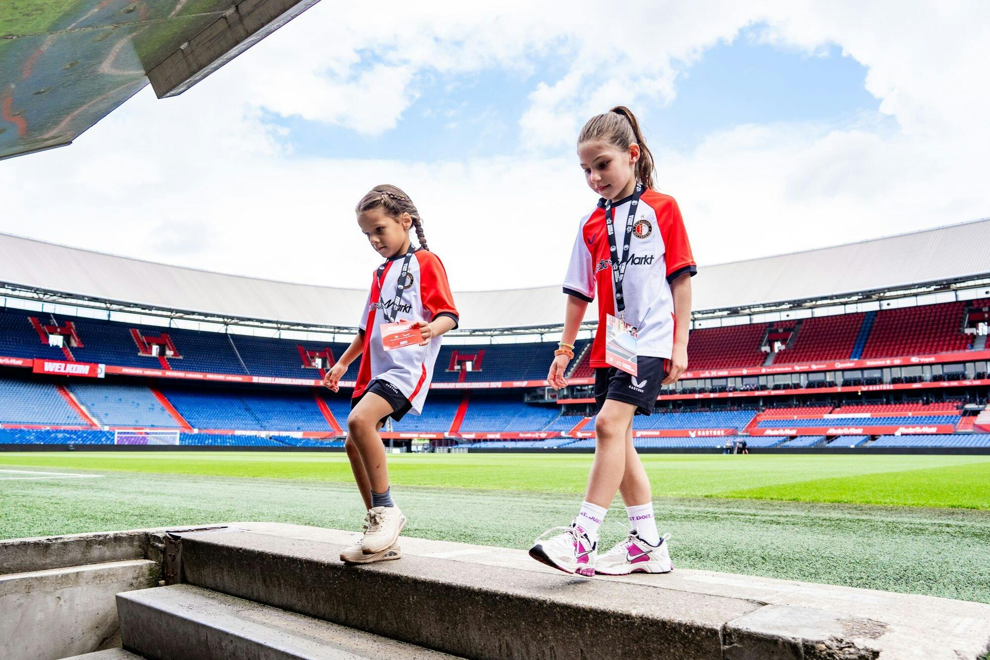 Duas crianças com camisolas desportivas vermelhas e brancas descem as escadas de um grande estádio com lugares vazios.