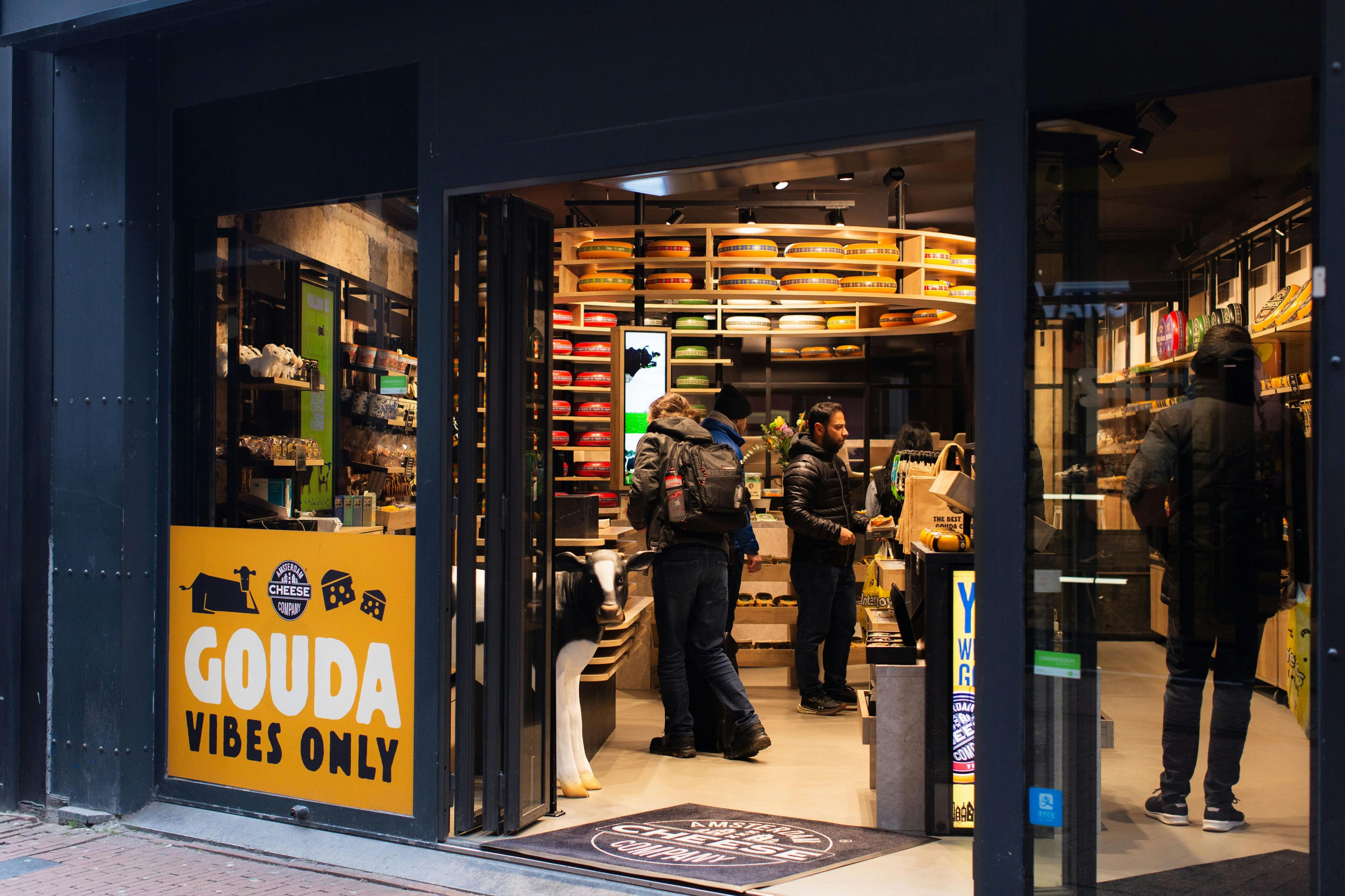 En ostbutiksinteriör med hyllor av osthjul och kunder som surfar. På en skylt utanför står det ”Gouda Vibes Only”.