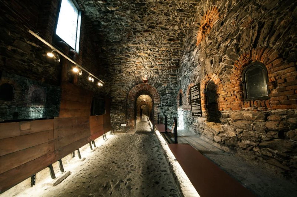 A stone-walled interior corridor with arched doorways, lit by spotlights, and a rough cobblestone floor.