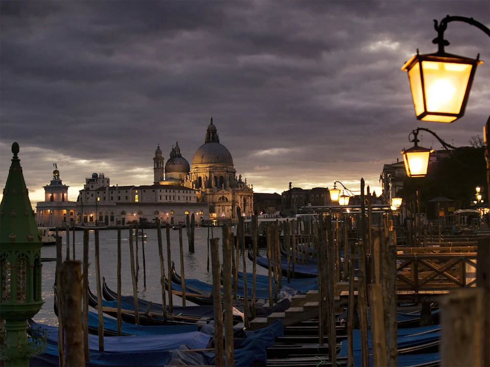 Gondolas moored at dusk in Venice, with street lamps illuminating the scene and a cathedral in the background under a cloudy sky.