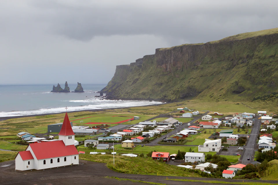 Small coastal town with a red-roofed church, scattered houses, cliffs, and sea stacks under an overcast sky.