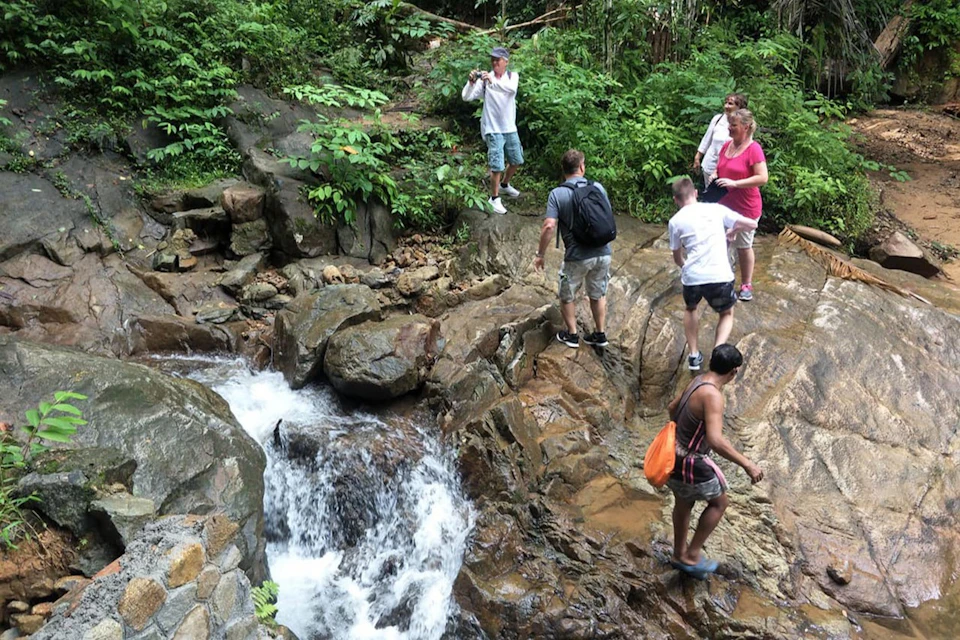 People explore rocky terrain near a small waterfall surrounded by lush greenery; one person is taking a photo.