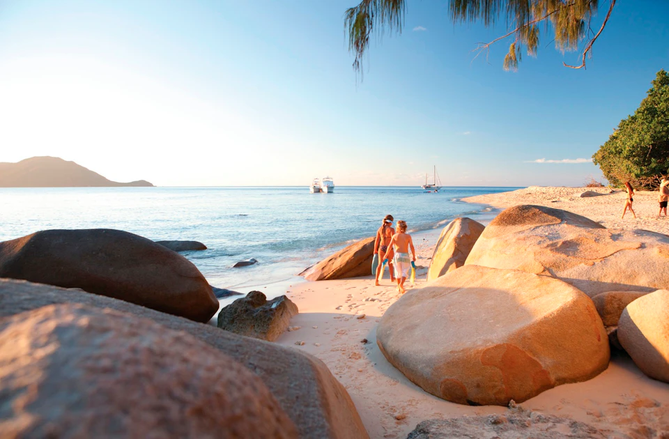 A couple walks on a sandy beach surrounded by large rocks, with boats in the distance under a clear blue sky.