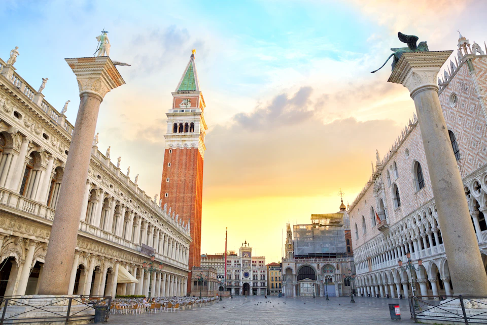 An empty plaza at sunrise features St. Mark's Campanile, arcades, and historic buildings in Venice.