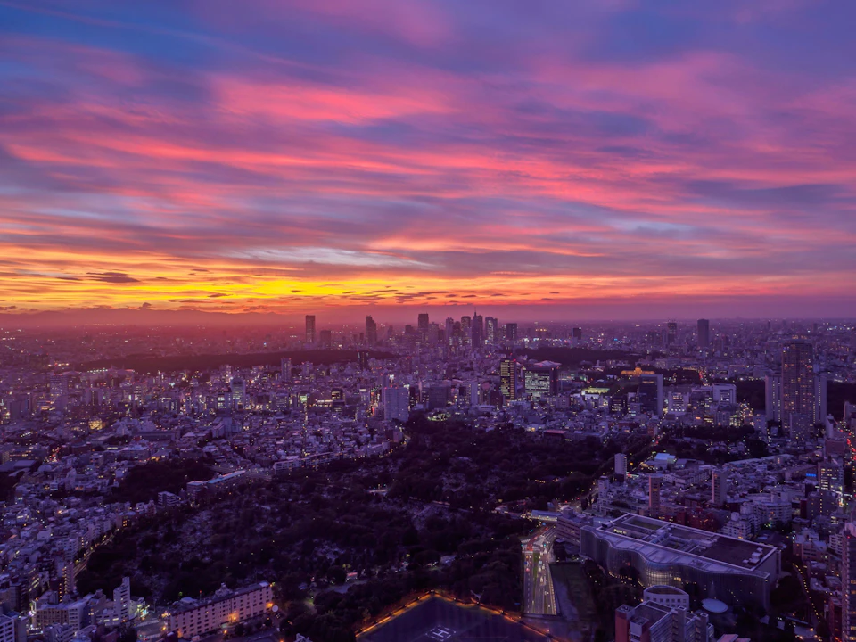 Cityscape at dusk with a vibrant sunset sky, showing illuminated buildings, streets, and a park in the foreground.