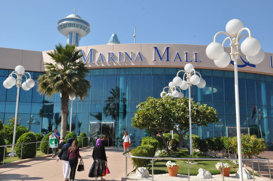 People walk towards the entrance of Marina Mall, with a glass facade, palm trees, and modern street lamps in front.