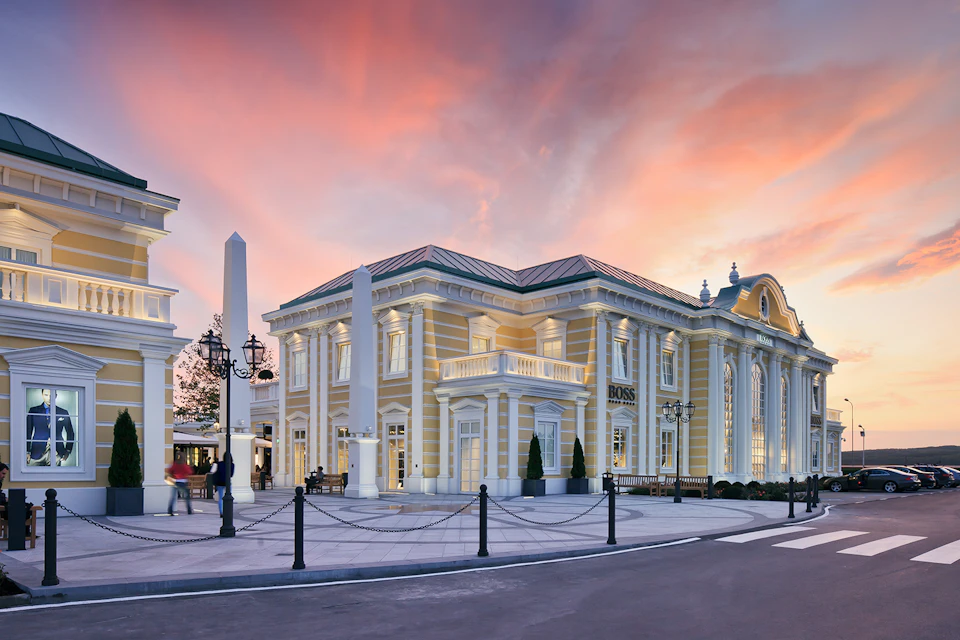 Elegant building with classical architecture features large windows and columns under a vibrant, sunset sky. Small statues and a few people nearby.