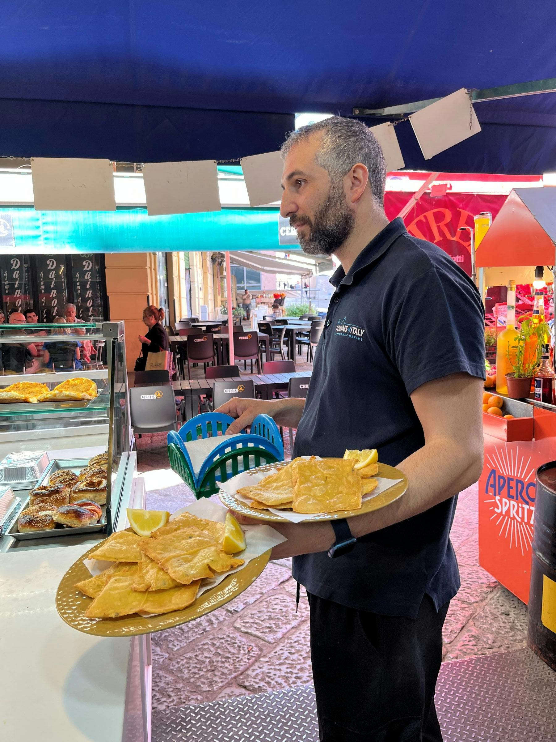 A man holding plates of food at an outdoor food stall with a blue canopy and a vibrant street market in the background.