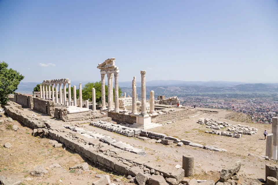 Ancient stone ruins with tall pillars and partial structures on a hilltop, overlooking a vast landscape under a clear blue sky.