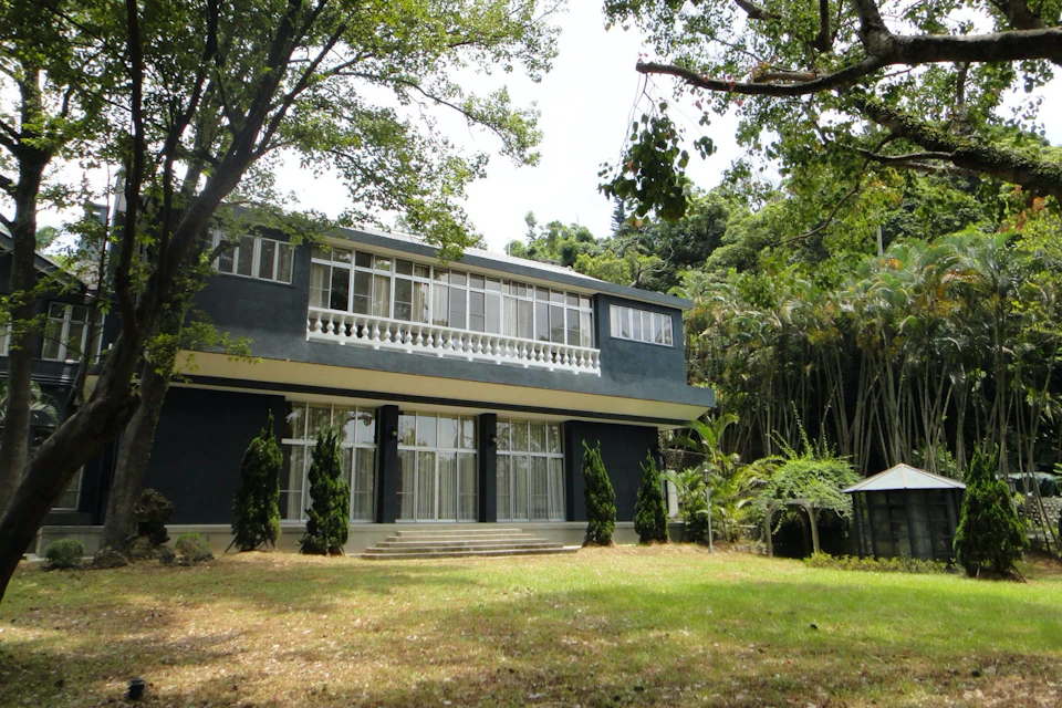 A two-story building with large windows and a balcony, surrounded by trees and greenery on a sunny day.