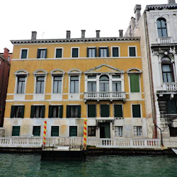 Tre angränsande byggnader med en kanal framför. Den centrala gula byggnaden har välvda fönster och gröna fönsterluckor.