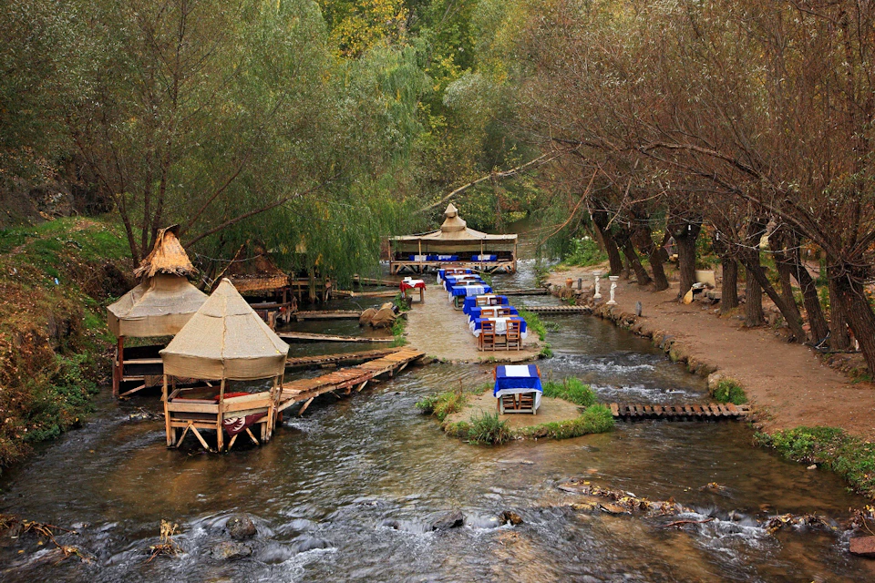 A dining area with blue-covered tables and chairs set on platforms over a creek, surrounded by trees and thatched huts.